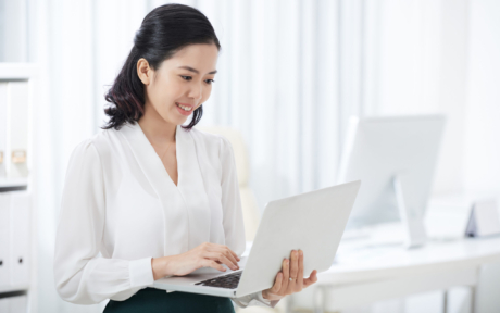 Beautiful young Asian woman in elegant outfit holding and using laptop with smile in modern office